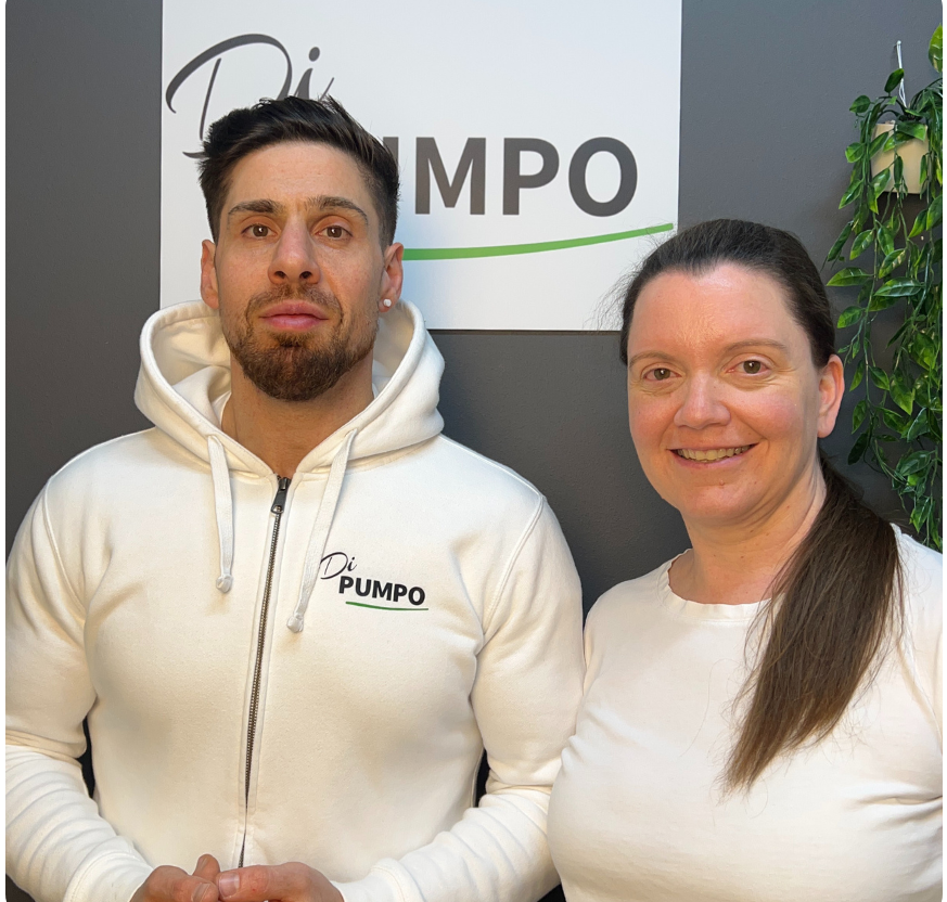 Zwei Menschen stehen in Werdohl vor einem Firmenlogo, lächeln und tragen sportliche Kleidung.