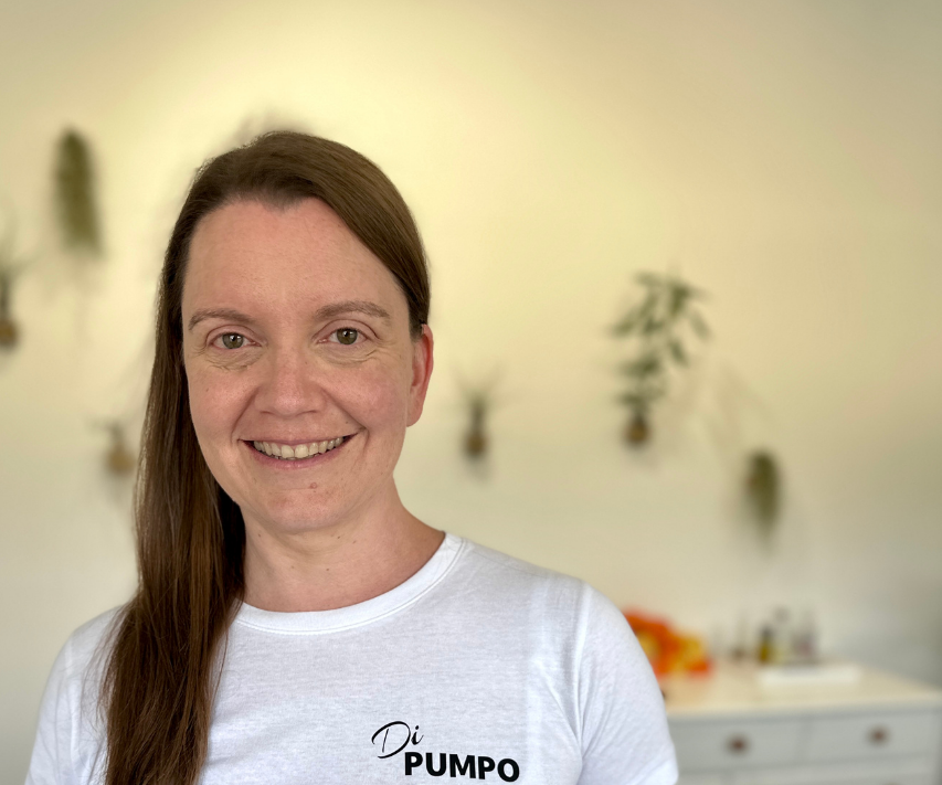 Frau mit langen braunen Haaren, lächelnd in einem hellen Raum mit Pflanzen im Hintergrund.